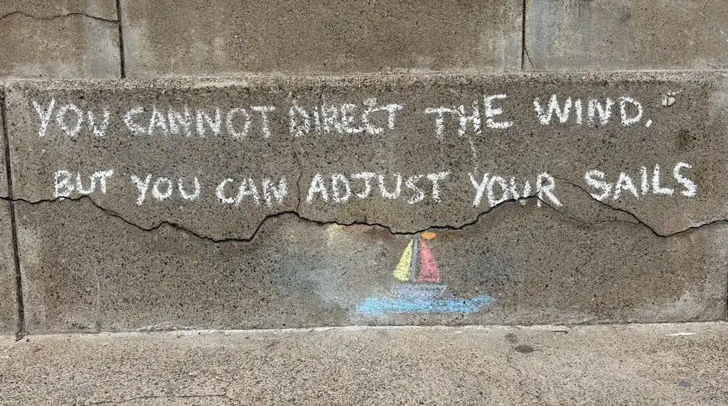 Chalk writing on a concrete step: You cannot direct the wind, but you can adjust your sails, above a small drawn sailing boat.
